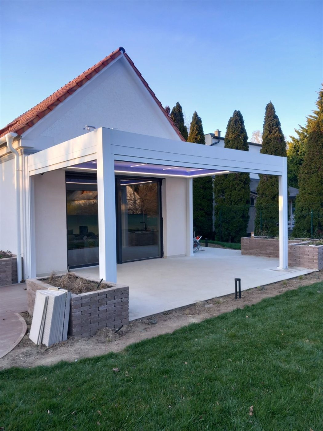 Fehér keretes pergola lamellavilágítással, kerti terasz alkonyatban